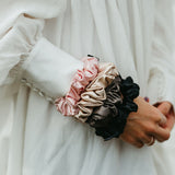 Stora sidensnoddar scrunchies i beige, svart, rosa, mörkgrå på en handled på kvinna med vit klänning
