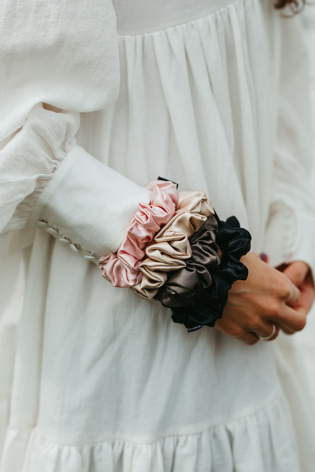 Stora sidensnoddar scrunchies i beige, svart, rosa, mörkgrå på en handled på kvinna med vit klänning